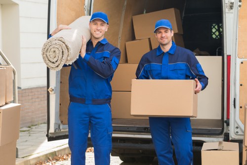 Two removal service workers dressed in blue uniforms are standing at the rear of a moving van parked outside a residential property in a suburban area. The worker on the left is smiling and holding a rolled-up beige carpet or rug over his shoulder with his right hand, while the worker on the right is also smiling and holding a medium-sized plain cardboard box in front of him. The interior of the van is visible, loaded with large cardboard boxes, some stacked and others loose, along with visible furniture pieces such as a wooden table, chairs, and a wardrobe, all wrapped for transport. The setting includes a driveway with a brick wall and parts of a house in the background under diffuse natural daylight. The scene captures the process of house removals, highlighting the careful handling of household items during a relocation, and subtly reflects services offered by Surrey Man with Van in the Surrey area, focusing on efficient and professional moving support.