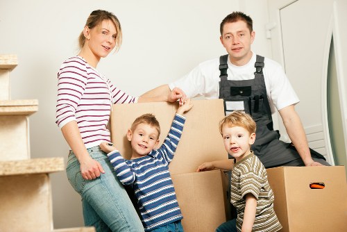 A family of four, including a woman with blonde hair wearing a red and white striped top and jeans, a man dressed in a white shirt and black work overalls, and two young boys in striped shirts, are indoors preparing for a home move. The woman and the boy in the blue and white striped shirt are holding a large cardboard box together, with the boy smiling at the camera. The other boy, in a beige and brown striped shirt, stands nearby with a slightly curious expression. The father is kneeling on the right, gently holding a smaller cardboard box, while the mother stands on the left at the bottom of a staircase with beige carpeted steps, in a residential interior with white walls and a door visible in the background. The scene depicts a household in the process of packing or moving, highlighting the personal belongings and family involvement typical of house removals in Surrey, near the postcode area. The boxes are plain, brown cardboard, and the overall lighting is bright and natural, emphasizing a calm, organized atmosphere associated with professional removals and sustainable home relocation services.