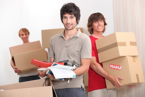 The image shows three young men in a bright, modern residential interior, likely a hallway or living room, preparing for a house removal. The man in the foreground has dark, wavy hair and a light grey shirt, holding a red clipboard, some papers, and a small black device, possibly a mobile phone or remote. Behind him, one man with blond hair and a light-colored shirt is carrying a medium-sized, plain cardboard box, while another man with medium-length brown hair and a red t-shirt is holding two stacked cardboard boxes, one labeled 'FRAGILE' with red and white tape. The background features light-colored walls and a window with sheer curtains, suggesting an indoor setting in Surrey or nearby town. All three appear to be engaged in packing or organizing, symbolising professional house removals services. The scene emphasizes household items such as cardboard boxes, with the focus on the preparation phase of moving home, consistent with Surrey Man with Van’s removal service activities in the local area.