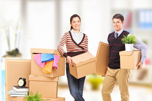 A man and a woman stand inside a bright, modern residential space, each holding cardboard boxes as part of a house move. The woman, on the left, has long dark hair and is smiling, wearing a striped sweater with shades of red, white, and grey, paired with jeans. She is holding a medium-sized cardboard box in her hands. Next to her are several stacked moving boxes, some wrapped with colorful cloths or blankets, which are placed on a clean surface. The man, on the right, has short dark hair and is dressed in a dark sweater with a striped pattern over a light shirt, beige trousers, and is smiling while holding two smaller boxes: one with a small potted green plant on top. The background is blurred but suggests a contemporary household with large windows and light-filled rooms, indicating that the scene takes place indoors during a house removal. The overall setting emphasizes professional house removals services in Surrey, with a focus on careful handling of household belongings and professional movers, consistent with Surrey Man with Van’s offerings in the local area.