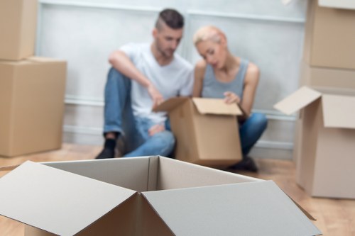 A man and woman are seated on the floor of a residential interior surrounded by several cardboard moving boxes, with the woman holding an open box and both appearing to be packing or unpacking belongings. The woman has short blonde hair and is wearing a light gray sleeveless top, while the man has dark short hair and is wearing a white T-shirt and blue jeans. In the foreground, an open cardboard box is empty, positioned centrally in the image. The background includes a plain light-colored wall and a few more boxes stacked on a wooden floor, suggesting an ongoing house move within a suburban or residential setting. The overall scene conveys preparations for a house relocation, with the individuals actively handling household items, typical of professional removals services such as Surrey Man with Van operating in the local area near Surrey or the South East UK postcode region. The lighting appears natural, illuminating the workspace as they organize contents in a typical home environment.