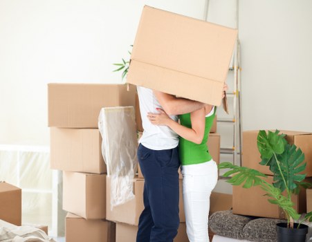 In a well-lit, neutral-toned room within a residential property, two movers from Surrey Man with Van are engaged in a house removal. One person, dressed in a white t-shirt and dark blue trousers, is holding a medium-sized cardboard box with a smooth, brown surface, positioned over their shoulder. The other person, wearing a bright green top and white trousers, appears to be assisting or receiving the box. Several other cardboard boxes, some sealed and others open with packing paper inside, are stacked around them, indicating an ongoing packing or unpacking process. To the right, a potted plant with large, green, glossy leaves is situated on a patterned cushion or small stool, adding a touch of greenery and natural texture to the scene. The background features a simple, light-colored wall with minimal decoration, emphasizing a typical home interior in the Surrey area, possibly within a living room or hallway. The ambient lighting suggests daytime, with natural light illuminating the space, supporting the safety and care focus of the removals service while visually grounding the scene in a UK household context.