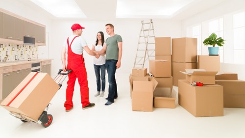 In a bright, spacious room with large windows allowing natural light, a man in a grey T-shirt and dark jeans is shaking hands with a mover dressed in red overalls, a white shirt, and a red cap. Behind them, a woman in casual clothing stands observing the interaction. The room contains a metal ladder leaning against the wall, with a potted plant on a windowsill to the right. Several cardboard boxes of various sizes are stacked and scattered around, some open revealing packing materials, while others are sealed. A hand truck loaded with a large package is visible on the left side of the image. The setting suggests an ongoing house move, with new or packed belongings awaiting transport. The interior appears clean and modern, indicating a residential property within the Surrey area, possibly related to the services offered by Surrey Man with Van, a professional removals provider specializing in local house relocations within Surrey postcode districts like GU or KT.