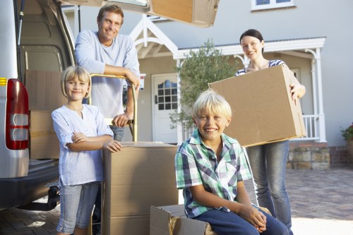 A family of four, consisting of a father, mother, and two children, are positioned outdoors near a grey removals van parked in a residential driveway. The father, dressed in a light blue shirt, stands at the back of the van, resting his arms on the open trunk, while the mother, wearing a striped top, holds one side of a large cardboard box. The two children, a girl and a boy with blond hair, are engaged in the moving process; the girl, in a light blue shirt, stands to the left, smiling with her arms crossed, and the boy, wearing a checkered shirt, is seated on a closed cardboard box, also smiling. The background features a light-colored detached house with a porch and a garden, set under a clear sky. The scene captures a moment of unpacking or packing during a house move, with visible household items and cardboard boxes highlighting the process, in a suburban neighbourhood typical of the Surrey area, supporting local house removal services like Surrey Man with Van.