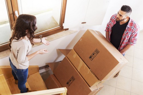A man and a woman are indoors at the entrance of a residential property, involved in a house removal process. The woman, with long brown hair and wearing a light-colored sweater and dark jeans, is standing on a wooden staircase, gesturing towards the boxes with her hands. The man, with dark hair and a beard, dressed in a red plaid shirt over a black T-shirt and dark trousers, is carrying a large cardboard box that appears to contain household items. They are positioned next to each other near the indoor doorway, with a white door and a window with a beige blind visible in the background. The staircase has a wooden handrail, and several large cardboard boxes are stacked at the bottom of the stairs, likely being prepared for relocation. The lighting appears natural, and the environment is clean and well-lit, reflecting a typical residential setting in the Surrey area. This scene illustrates the process of household item packing and moving, related to professional removals services offered by Surrey Man with Van.
