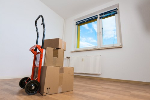 The image shows a minimally furnished room with white walls and light wooden flooring, illuminated by natural daylight coming through a large, double-glazed window with blue and yellow window trims. In the foreground, there are three cardboard boxes of varying sizes, stacked with two large boxes at the bottom and a smaller one on top, all appearing to be ready for moving or recycling. An orange hand truck with black handles and wheels is positioned next to the boxes, leaning against the wall. The room contains a white radiator beneath the window, and no other furniture or personal belongings are visible. The scene reflects a typical interior space in the process of being cleared or prepared for a house move, with the presence of packing materials and equipment associated with professional removals services, such as Surrey Man with Van, operating in the Surrey or nearby areas. The overall atmosphere suggests a clean, organized environment suitable for house relocations or clearing projects related to sustainable moving practices within the local community.