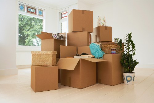 A collection of cardboard moving boxes of various sizes and shapes are stacked in a bright, spacious room with light wooden flooring. Some boxes are sealed with packing tape, while others are open, revealing contents such as fabric items and household objects. A woven storage box and a smaller cardboard box are placed on the floor among the stacks. A turquoise cloth covers part of one box, and a wicker basket with more items is also visible. To the right, a tall potted plant with lush green fern-like leaves in a decorative white and blue ceramic pot adds a natural element to the scene. In the background, large windows allow natural light to fill the room and offer a view of green trees outside. The room appears empty of furniture, indicating it is in the process of being cleared or prepared for a move, typical of house removal activities. The scene suggests a house in the UK, possibly within Surrey, ready for a scheduled move managed by Surrey Man with Van, specializing in removals.