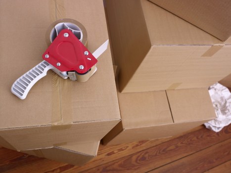 The image shows several sealed cardboard boxes stacked in a residential interior, likely part of a house removal or moving process. The boxes are made of standard brown corrugated cardboard with visible flaps and tape, some of which are taped shut, indicating packing for a move. On top of one of the boxes is a red and white toggle switch device, possibly a safety or electrical switch. The background includes a wooden floor with a natural finish, and the scene appears to be inside a house or apartment, possibly in Surrey, given the context of Surrey Man with Van's services. The lighting is natural, illuminating the area evenly, with the focus on the boxes and the switch device, which suggests careful packing and organization typical of a professional removals service. The scene portrays an organized moving environment, with household items prepared for transport, reflecting the company's focus on efficient and safe house relocations within the local area.