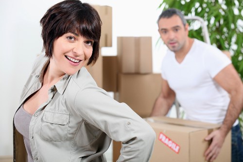 A woman with short dark hair, wearing a light grey shirt, is smiling and looking directly at the camera in the foreground of the image. Behind her, a man with short dark hair and a beard, dressed in a white t-shirt, is slightly out of focus as he carries a cardboard box. They are in a residential interior, likely a house undergoing a move, with several packed cardboard boxes stacked around them. Some boxes are sealed with tape and labeled, indicating they are packed for removal or relocation. The background features a welcoming home environment with a white wall, green plants, and a staircase or hallway leading further inside. The natural lighting illuminates the scene, highlighting the textures of the cardboard boxes, the man's white shirt, and the woman's attire. The scene captures a professional house removal process, closely associated with Surrey Man with Van’s services, demonstrating the careful handling of household belongings during a move in the Surrey area near the postcode for the town, subtly referencing local relocation services and safety procedures in home removals.