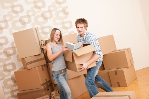 A young couple with light skin and casual clothing are engaged in packing or unpacking boxes during a house move inside a residential room, with several large cardboard boxes stacked against a wall with a patterned wallpaper in the background. The woman, dressed in a sleeveless top and jeans, holds a brochure or pamphlet, smiling as she interacts with the man. The man, wearing a checkered shirt and jeans, is holding an open box, possibly filled with household items, and appears to be lifting or positioning it. The floor is wooden, and additional boxes are placed on the floor, suggesting an ongoing moving process. The scene is well-lit, emphasizing the activity of loading or sorting belongings typical of house removal services in Surrey or nearby areas, with natural and ambient lighting creating a neutral, professional environment overall. This image effectively illustrates the process of residential relocation involving furniture, packing boxes, and teamwork, aligned with removals services offered by Surrey Man with Van.