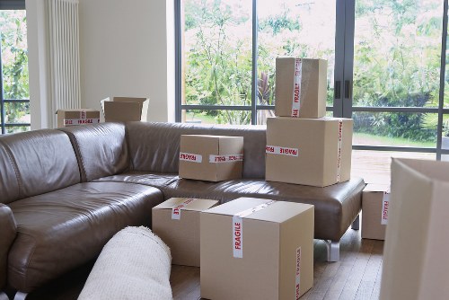 A living room interior with a large L-shaped brown leather sofa positioned against floor-to-ceiling windows, which reveal greenery outside, suggesting a house in Surrey. Multiple cardboard boxes, some labeled 'Fragile' with red tape, are placed on the sofa and on the wooden floor, indicating packing or unpacking activities. The boxes vary in size, with some stacked and others arranged individually, containing household items likely for a house removal. A white fabric cushion is seen on the sofa, adding a soft texture contrast. The natural light flowing through the windows illuminates the space, emphasizing the clean and organized environment typical of a house removal process. This scene subtly highlights Surrey Man with Van's removals services within a residential setting in the Surrey area, with a focus on detailed furniture and packing supplies essential for local house moves.