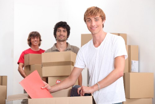 Three individuals involved in house removals or moving activities are indoors in what appears to be a residential setting, with cardboard boxes of various sizes stacked and arranged in the background. The person at the forefront, a young man with light brown hair wearing a white T-shirt, is holding a red folder while standing beside a partially filled cardboard box. Behind him are two other individuals, a woman with curly brown hair in a red top and a man with dark, slightly curly hair and a beard in a beige shirt, both smiling and holding packed boxes. The boxes are made of brown corrugated cardboard, some with white labels, and display different textures and sizes suitable for household items such as books, decor, or small appliances. The scene is well-lit with neutral lighting, emphasizing the domestic environment consistent with house removals or relocation services, subtly aligned with Surrey Man with Van’s service area near Surrey or South West London, and the setting suggests activity related to moving or packing. The environment appears clean and organized, highlighting professional packing and relocation tasks typical of house removal services in the local area.