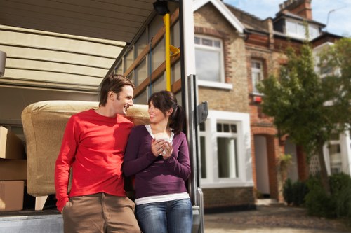 In the image, a young man and woman are standing outside the open rear of a removals van during the daytime, which is parked on a residential street in Surrey, UK. The man, dressed in a red long-sleeve shirt and beige trousers, is smiling and looking at the woman. The woman, wearing a purple jacket over a white top and blue jeans, is holding a clear glass and smiling back. Inside the van, visible in the foreground, are large cardboard boxes and a single large piece of beige upholstered furniture, possibly a armchair or sofa, with a textured fabric cover. The van's interior reveals an organized space for household items, with the boxes stacked neatly behind the furniture. In the background, semi-detached brick residential houses with white-framed windows and small front gardens lined with bushes can be seen. The scene appears to be a typical home moving situation during clear weather, with natural light illuminating the surroundings while both individuals convey a casual, friendly atmosphere associated with house removals and local moving services in Surrey.