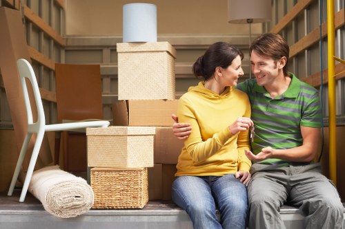 A man and woman sit closely together inside a storage unit filled with packed household items, including stacked cardboard boxes, woven baskets, a rolled-up rug, and a white plastic chair. The woman, wearing a yellow sweater and jeans, and the man, in a green and grey striped polo shirt and grey trousers, share a smile while looking at each other. Behind them, the background features wooden shelving supports and additional boxes, indicating a residential move or packing scenario typical in house removals and relocations within the Surrey area. The natural lighting highlights the textures of the cardboard, woven basket, and fabric clothing, realistically capturing the scene of careful packing and organization prior to transportation.