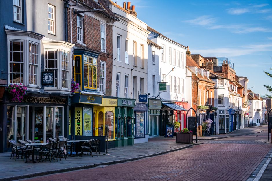 A narrow street in Guildford town centre during daytime, lined with small retail shops housed in historic brick and white-painted buildings. The shopfronts feature large glass windows displaying vario